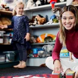 Smiling student sitting on the floor of a school playroom, with a young child in the background surrounded by toys