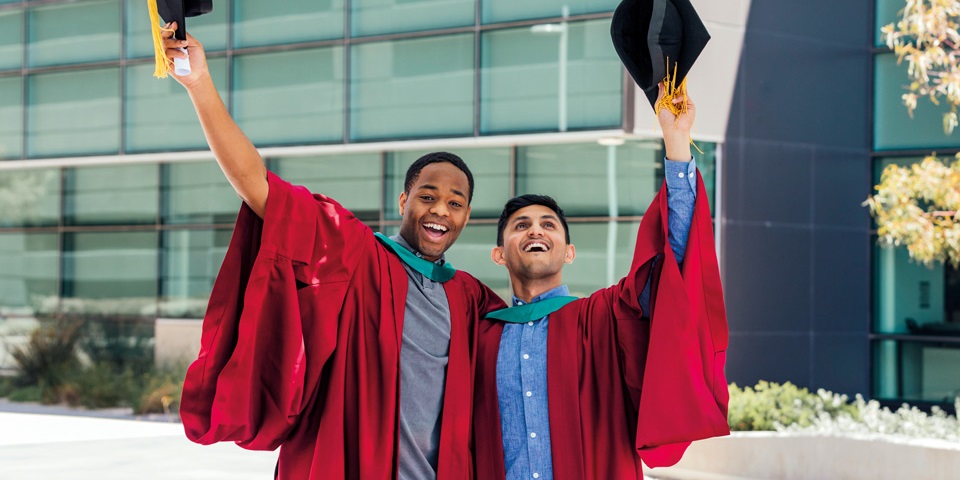 两名学生身穿毕业长袍，面带微笑，手拿帽子，庆祝毕业.