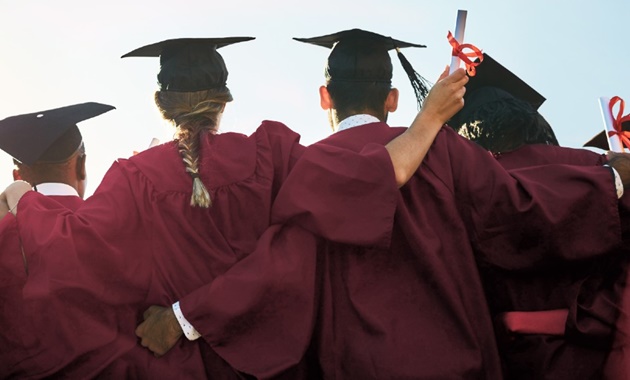 Four graduates in gowns and mortar boards standing with their arms around each other. Their backs are to the camera. Two graduates are holding diplomas tied with a red ribbon.