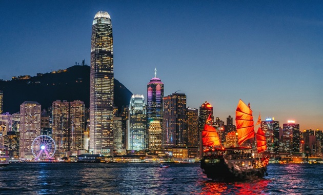 A red-sailed junk boat sailing in Hong Kong's Victoria Harbour at dusk, with the city in the background.