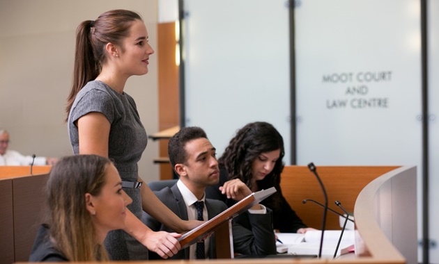 Female law student standing at Moot Court, with other students sitting either side.