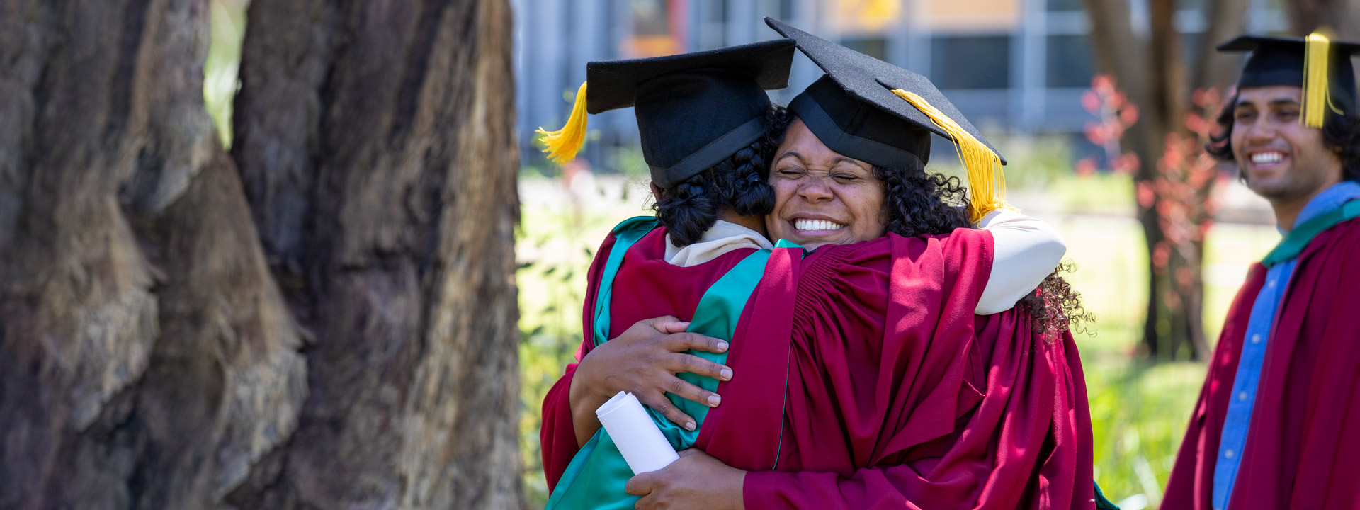 Two students smiling while hugging each other on graduation day. They are wearing their graduation robes and standing outdoors, next to a tree.