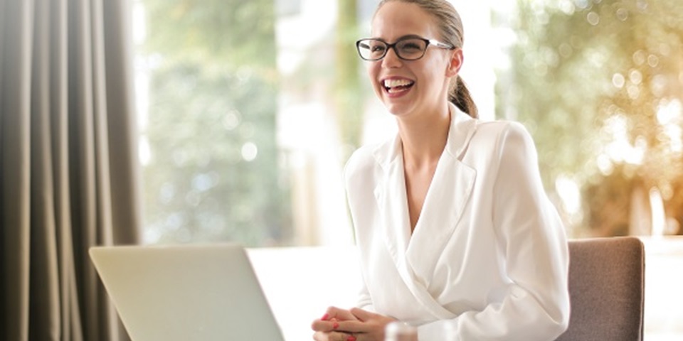 Woman in white blouse and glasses sits at a laptop, smiling.