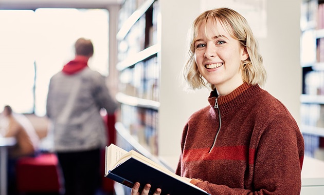 Female student in Library, holding a book, smiling at camera