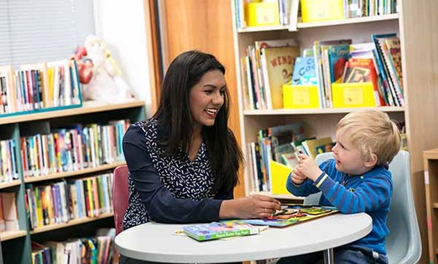 Teacher sits with early childhood student in library.