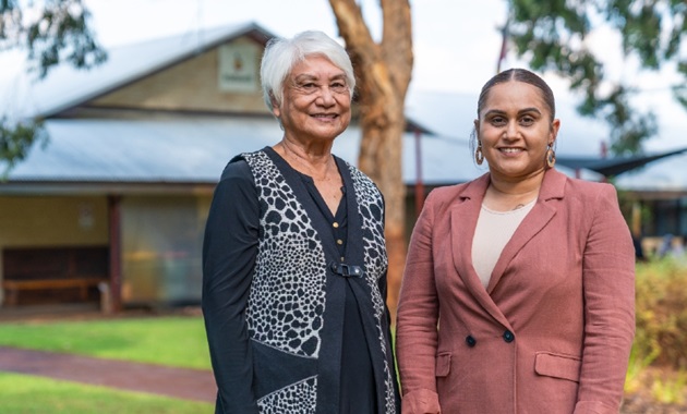 Two women smiling, looking at the camera, in front of a building shrouded in lush green native trees