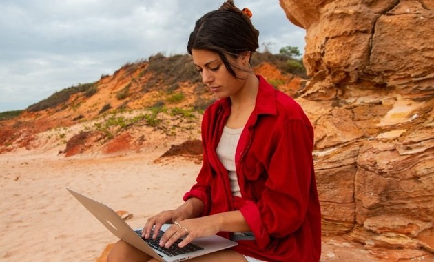 Student in red shirt sits on a red rock and studies laptop