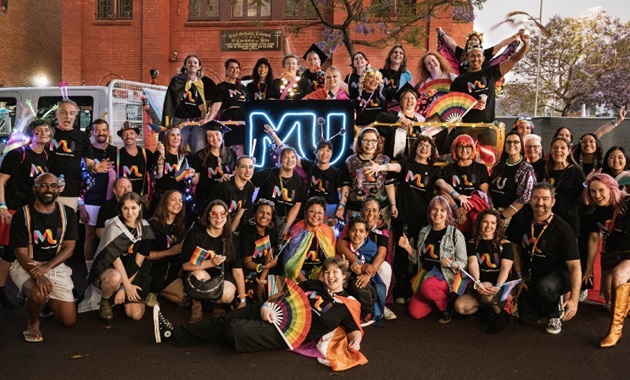 Large group of Murdoch University staff in black shirts with rainbow MU on front smiling at camera