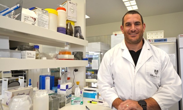 man with brown hair in a lab wearing a lab coat and smiling at the camera