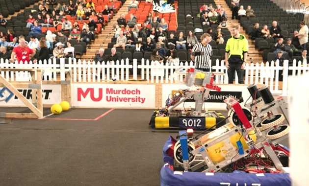 Two robots in the foreground in an indoor fenced area with stadium seats behind the short fence. Two people inside the fence with the robots and spectators in the stands.