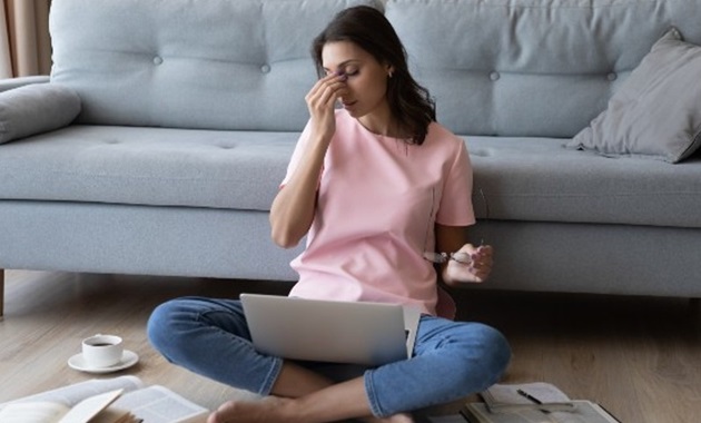 Young woman with books surrounding her as she sits on the floor with a laptop, holding her head.